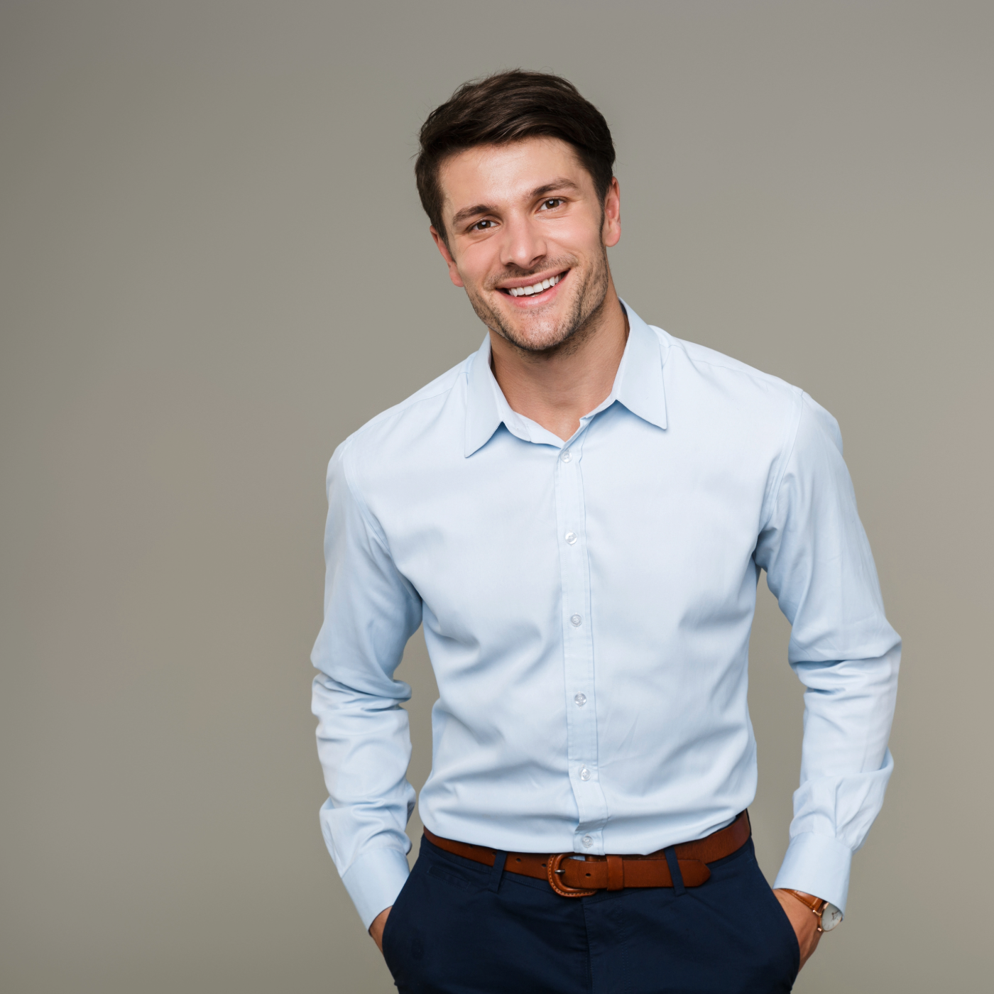Smiling young man wearing a light blue dress shirt and dark pants with hands in pockets against a plain background.