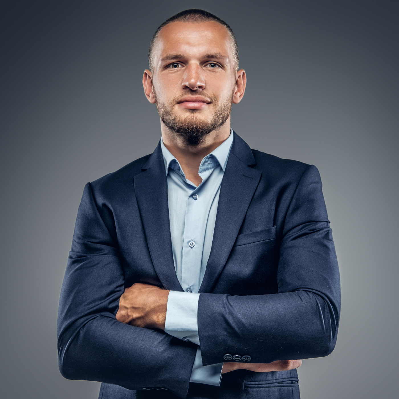 Confident man with short hair and beard in a dark blue suit and light blue shirt, arms crossed, against a gray background.