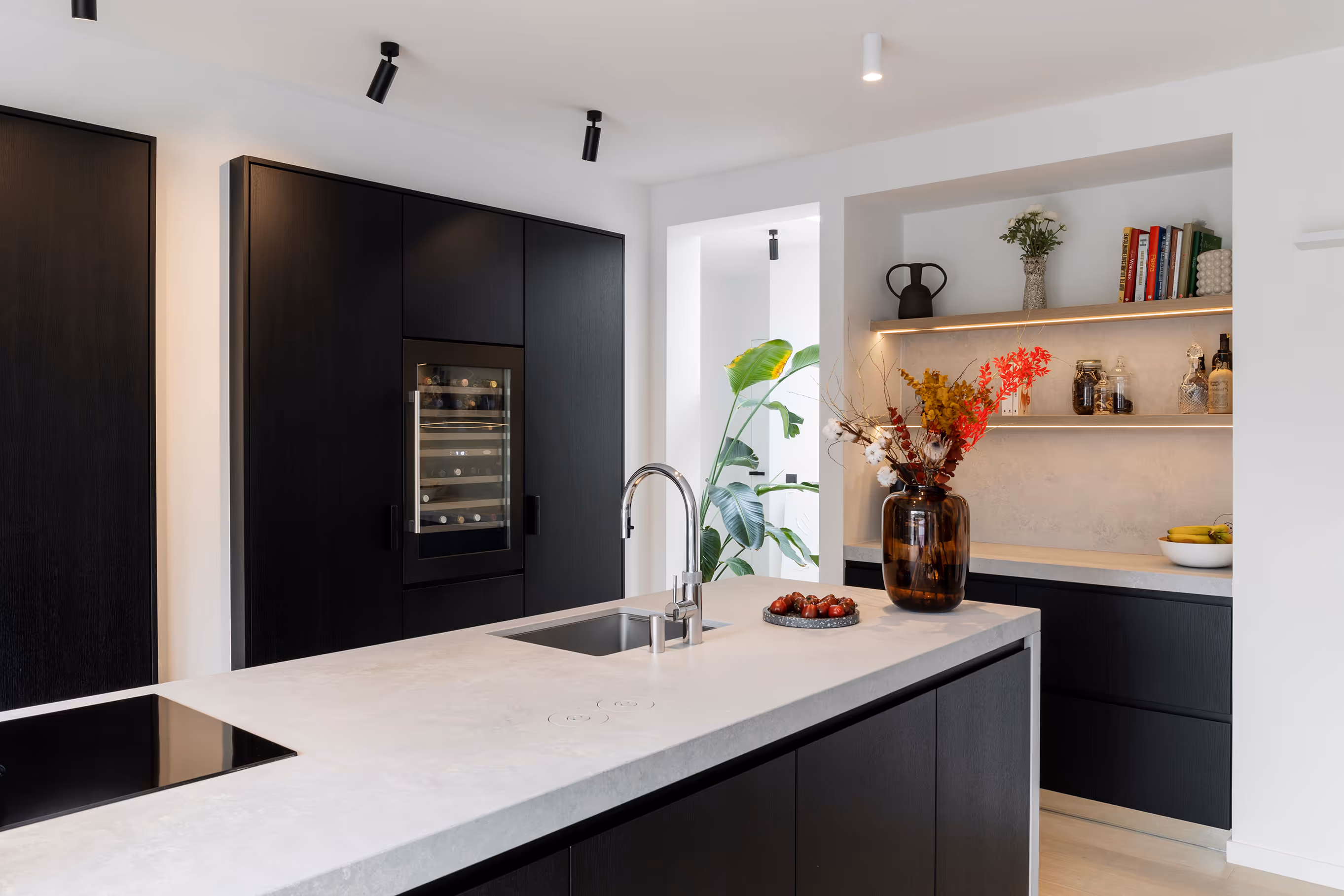 Modern kitchen with black cabinets, white countertop island with sink, vase with colorful flowers, and open shelves with books and decor.