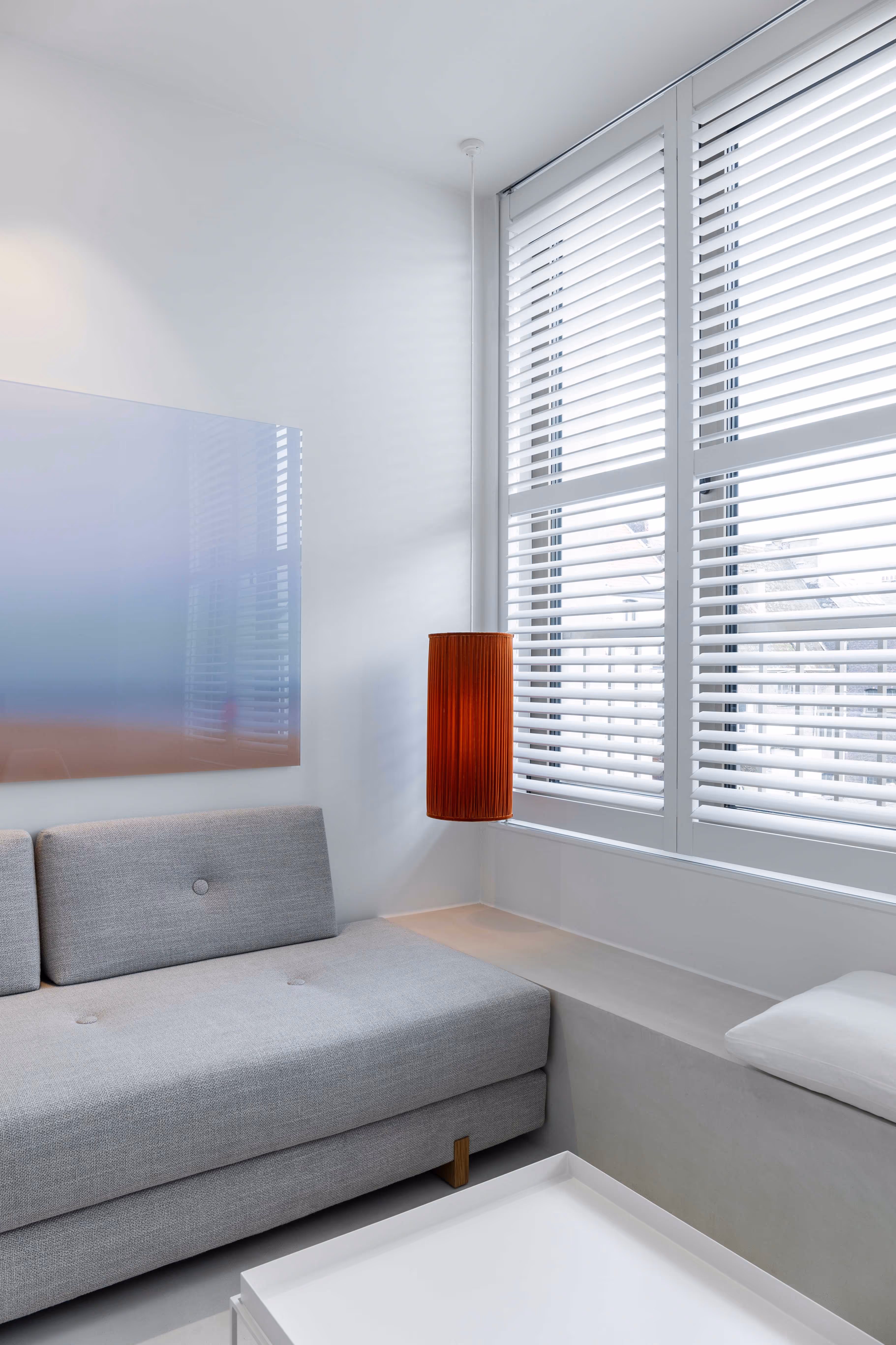 Modern living room corner with gray sofa, white window shutters, brown hanging lamp, and a white rectangular tray table.
