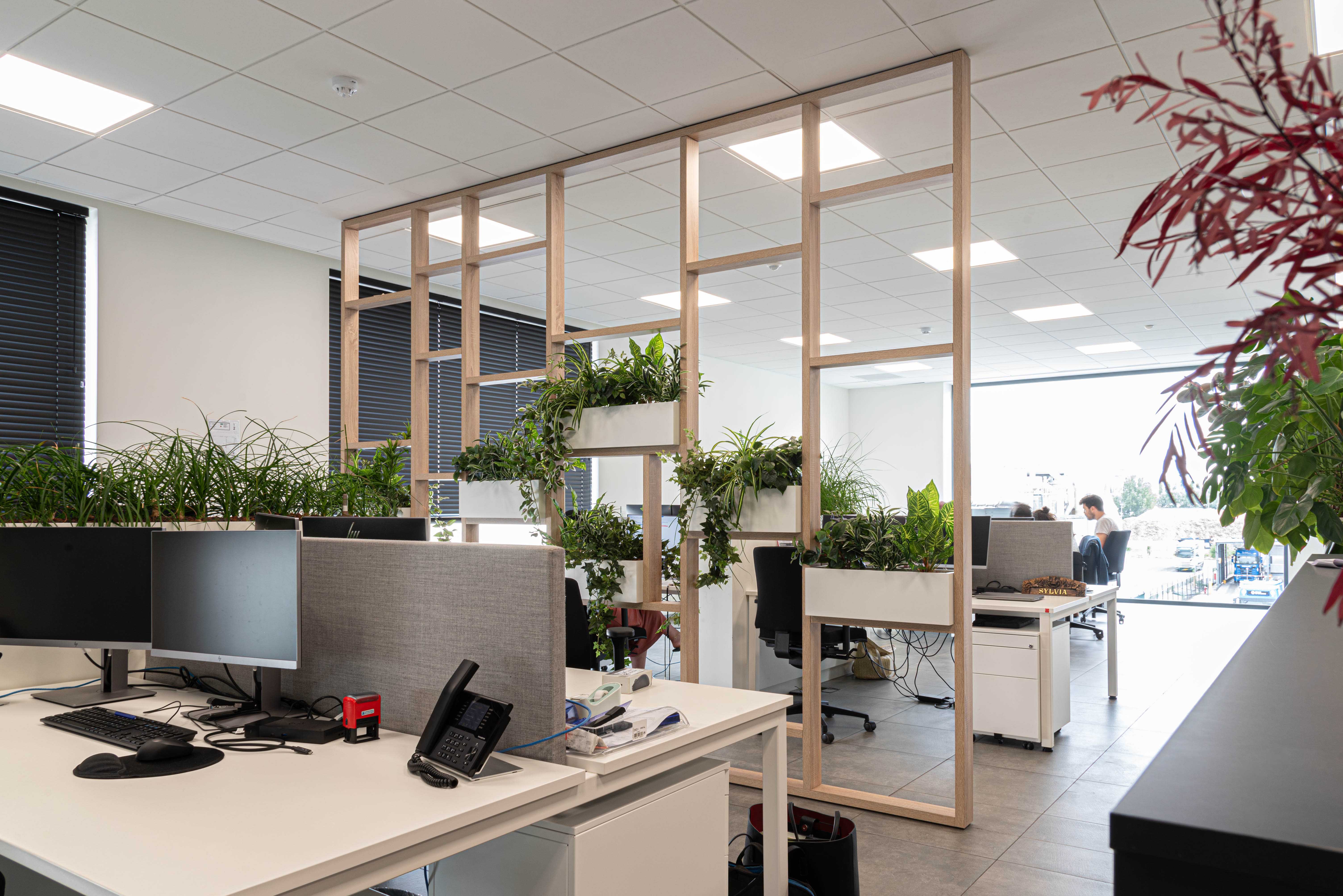 Modern office with white desks, computer monitors, a desk phone, and green plants integrated into wooden partitions and decor.