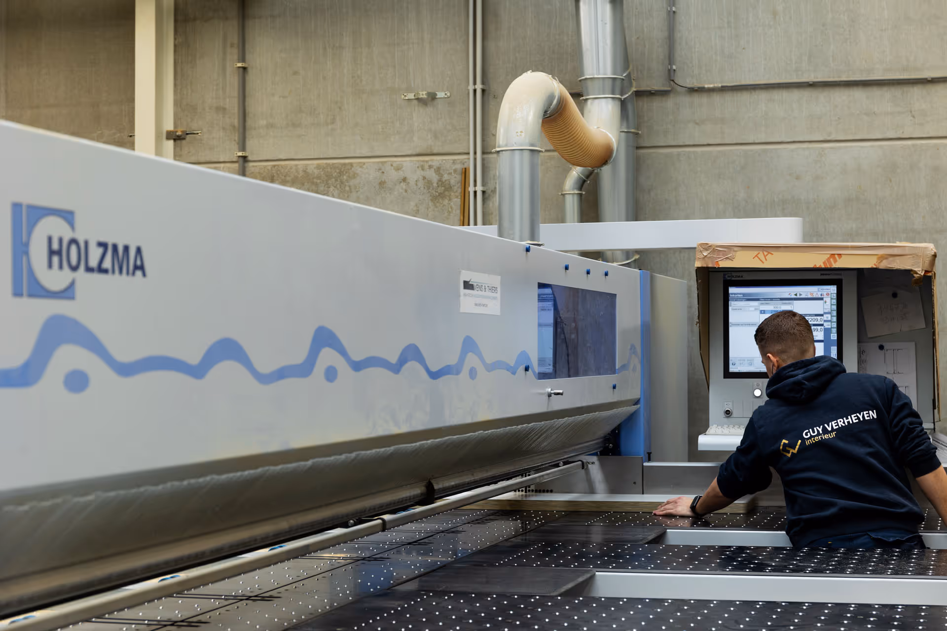 Man operating a large HOLZMA industrial woodworking machine with control screen in a workshop.