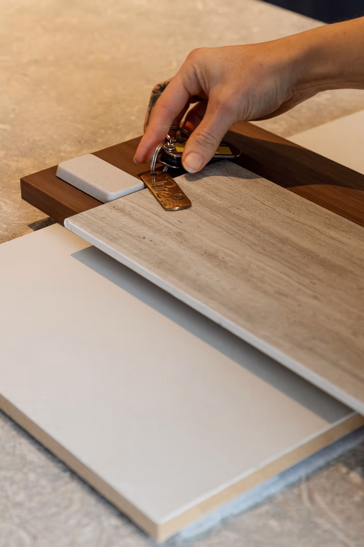 Hand holding a set of keys with a bronze tag over a selection of wood and tile samples on a table.