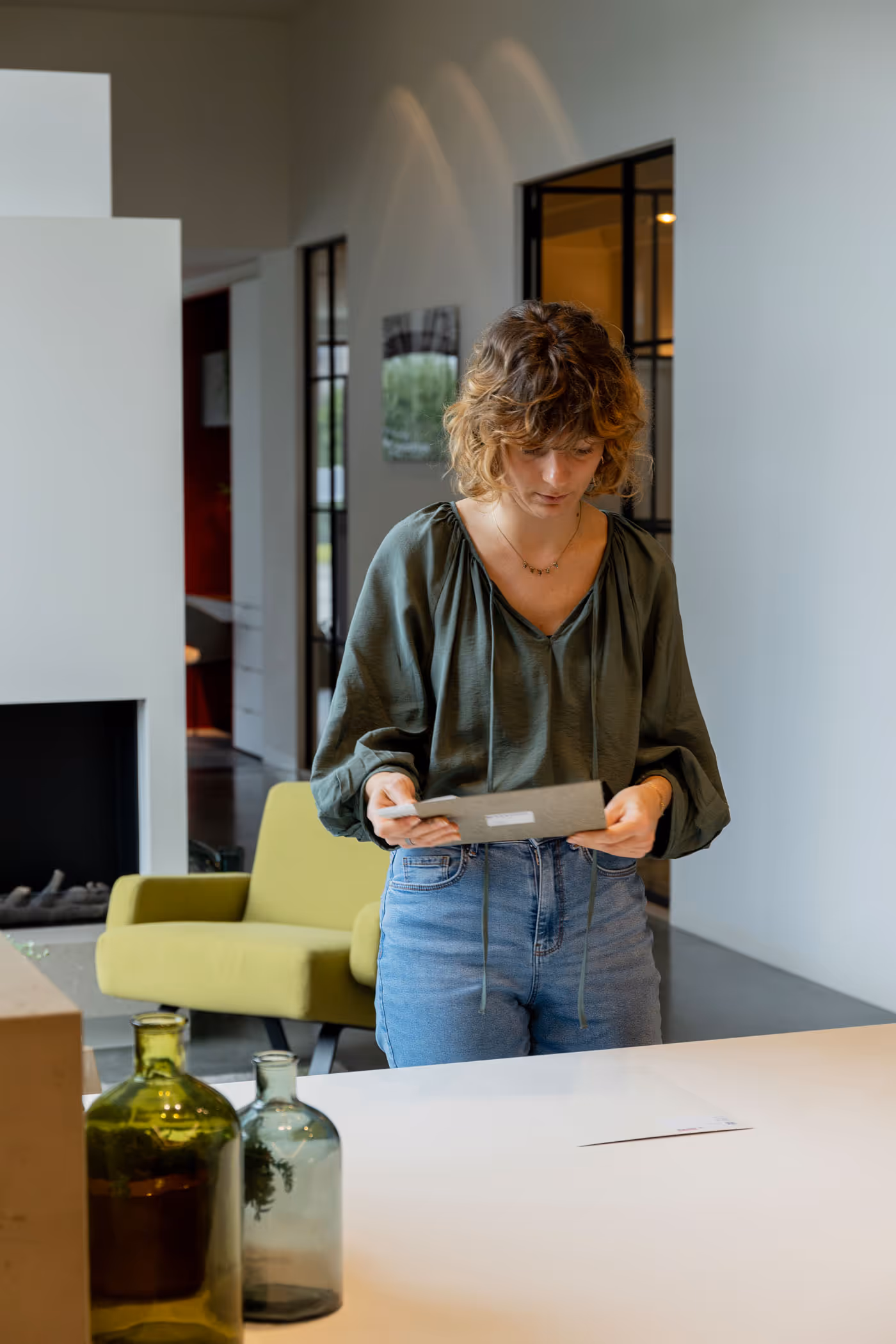 A woman in a green blouse and jeans standing indoors, reading a gray envelope, with green and clear glass bottles on a white table in the foreground.