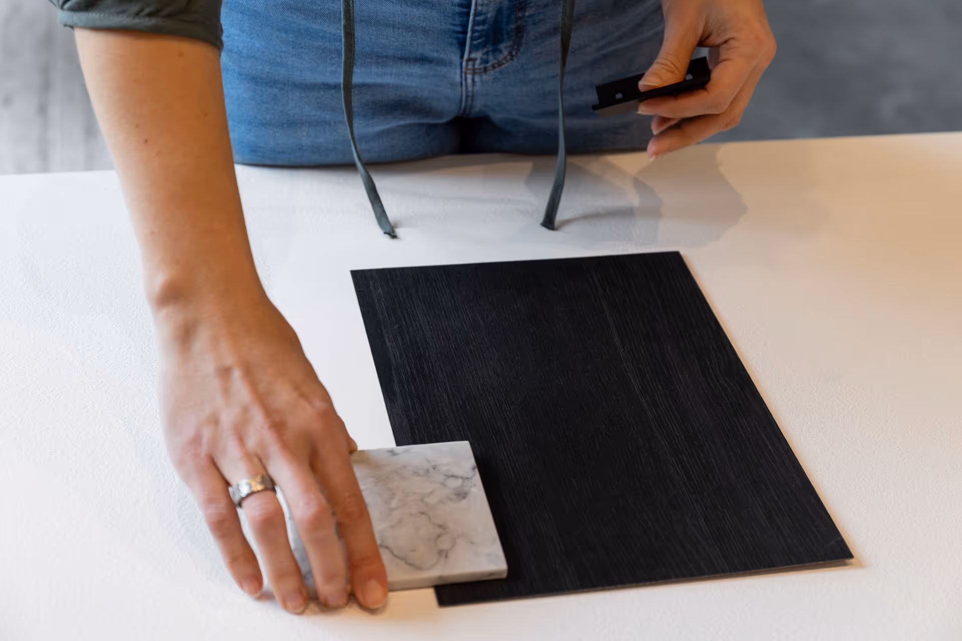 Person comparing a square black wood-textured panel and a small white marble tile on a white surface.
