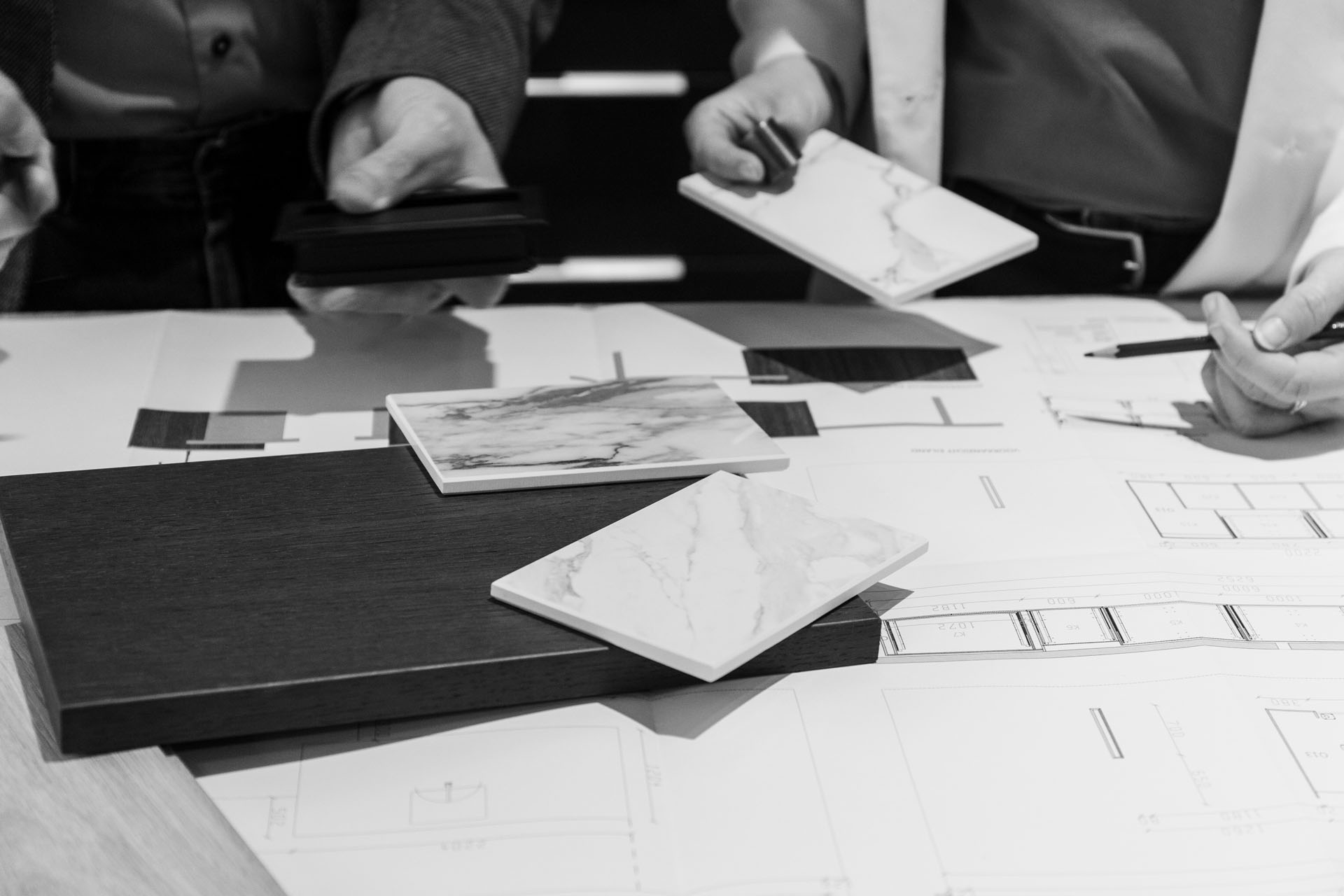 Two people holding laminate and tile samples over architectural blueprints on a table.