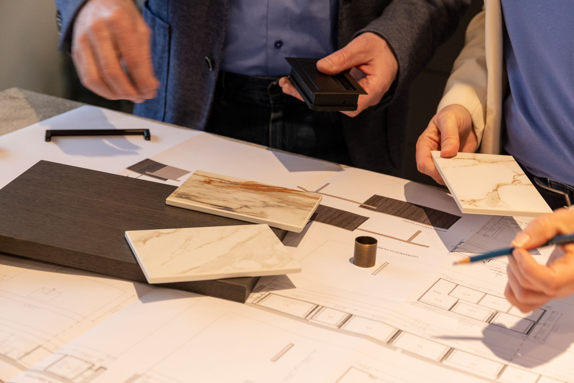Hands of two people examining marble tile samples, a dark wood sample, and a metal pull handle over architectural blueprints on a table.