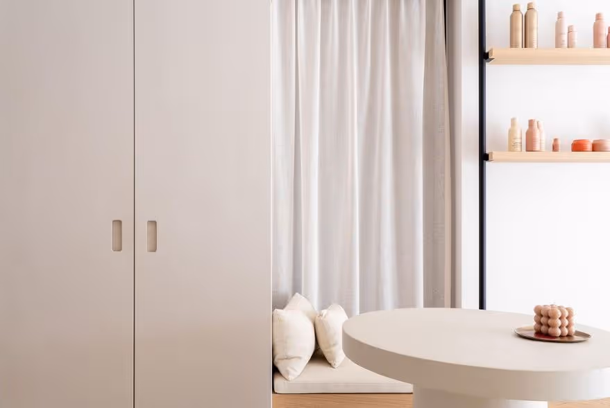 Minimalist beige interior with a round table holding a decorative candle, built-in shelves with bottles, and a cushioned seating area by white curtains.