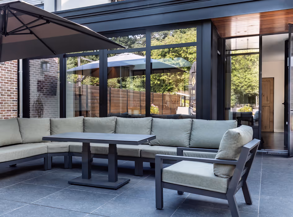 Modern outdoor patio with a gray cushioned L-shaped sofa, matching armchair, square table, and large umbrella with glass walls reflecting greenery.