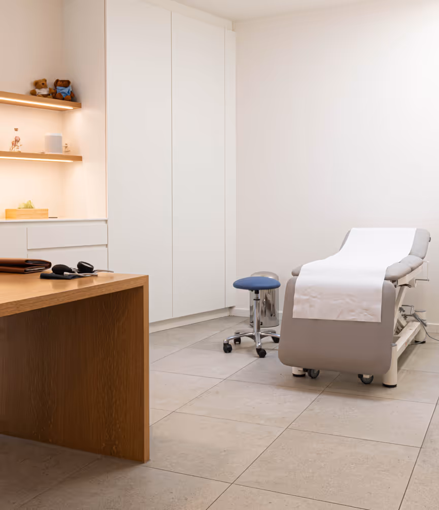 Minimalist medical exam room with a gray examination table covered in white paper, a blue rolling stool, wooden desk, and shelves with medical models and stuffed teddy bears.