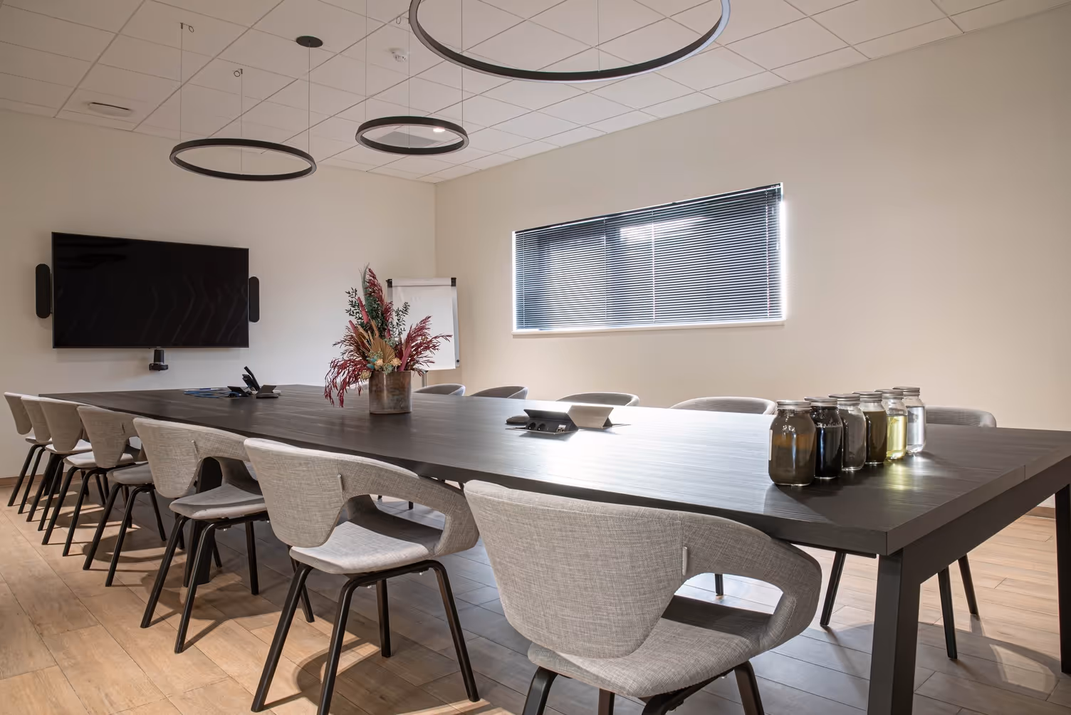 Modern conference room with a large dark wood table, beige chairs, circular ceiling lights, a TV screen on the wall, a window with blinds, and a vase of dried flowers as centerpiece.