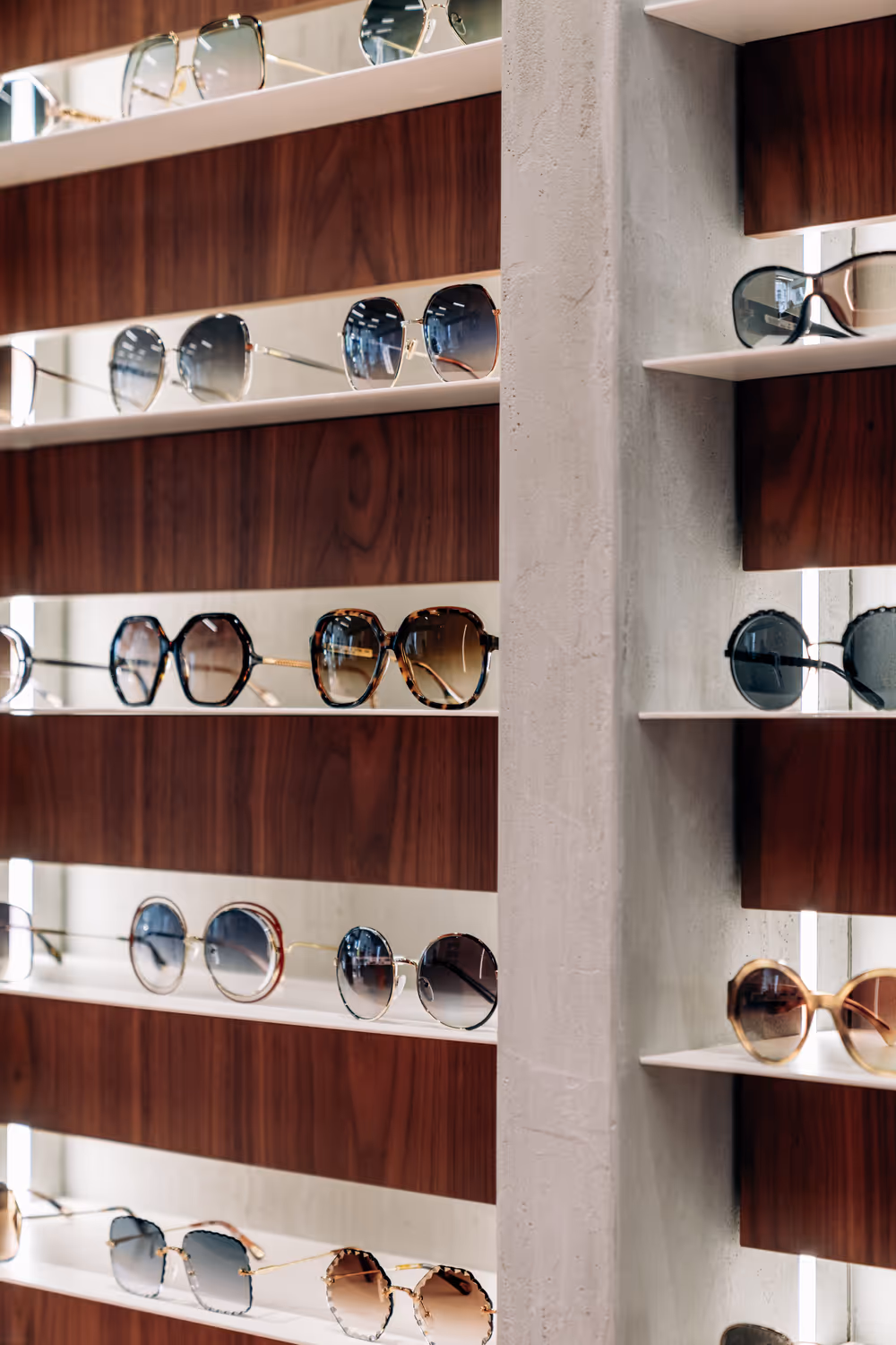 Display of various stylish sunglasses on white shelves with a wooden backdrop in an eyewear store.