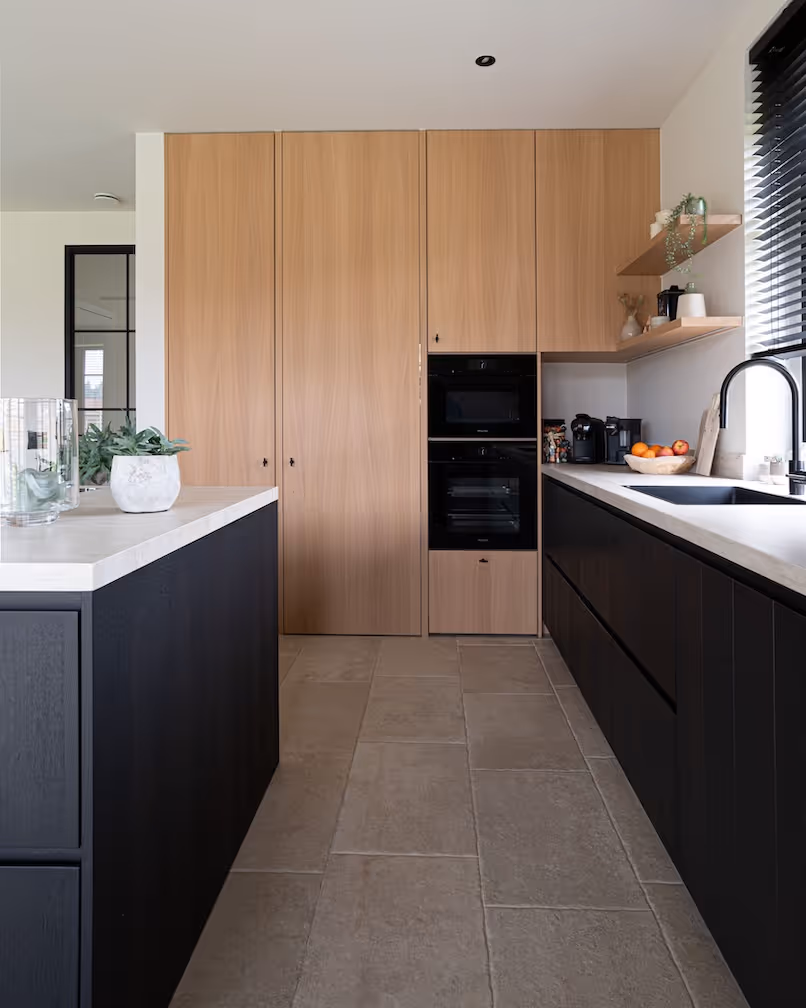Strakke keuken met zwarte onderkasten, lichte werkbladen en hoge kasten in warm houtfineer, met open houten wandplanken.
