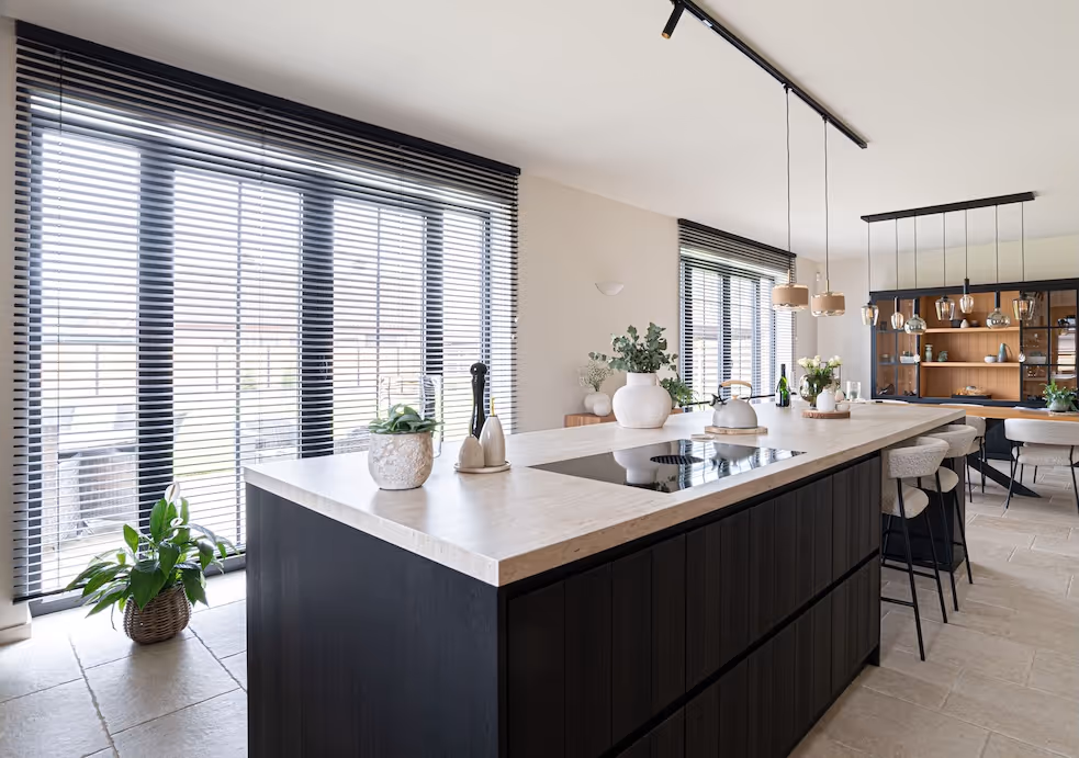 Lichte leefkeuken met donker kookeiland, natuurstenen werkblad en sfeervolle hanglampen, met zicht op een open eetruimte.