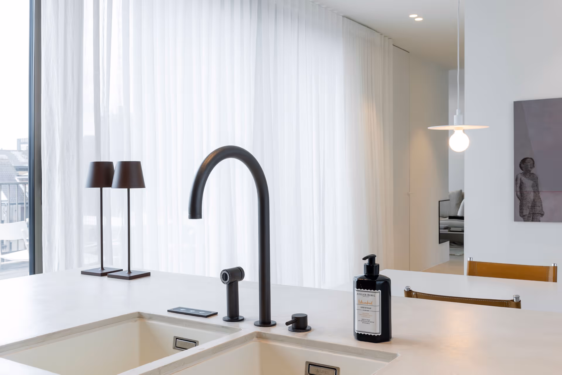 Minimalistische keuken met matzwarte kraan, dubbele spoelbak en designzeepdispenser, tegen een achtergrond van lichtgordijnen en moderne verlichting.