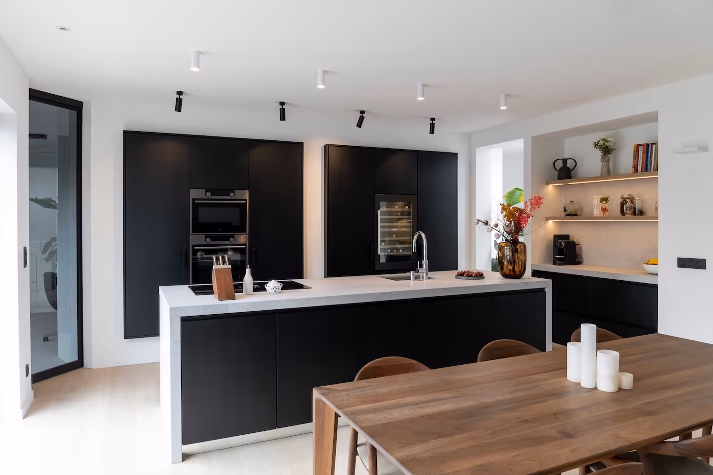 Moderne zwarte keuken met kookeiland, wijnklimaatkast en open wandrekken, naast een houten eettafel met kaarsen.