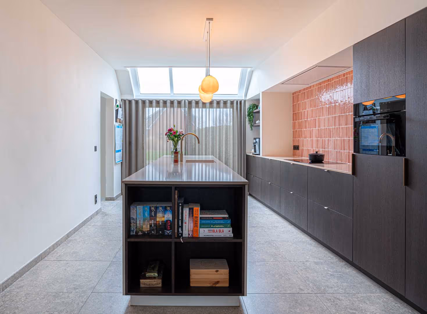 Moderne keuken met donker kookeiland, ingebouwde boekenkast, koperkleurige kraan en opvallende roze wandtegels onder een lichtkoepel.