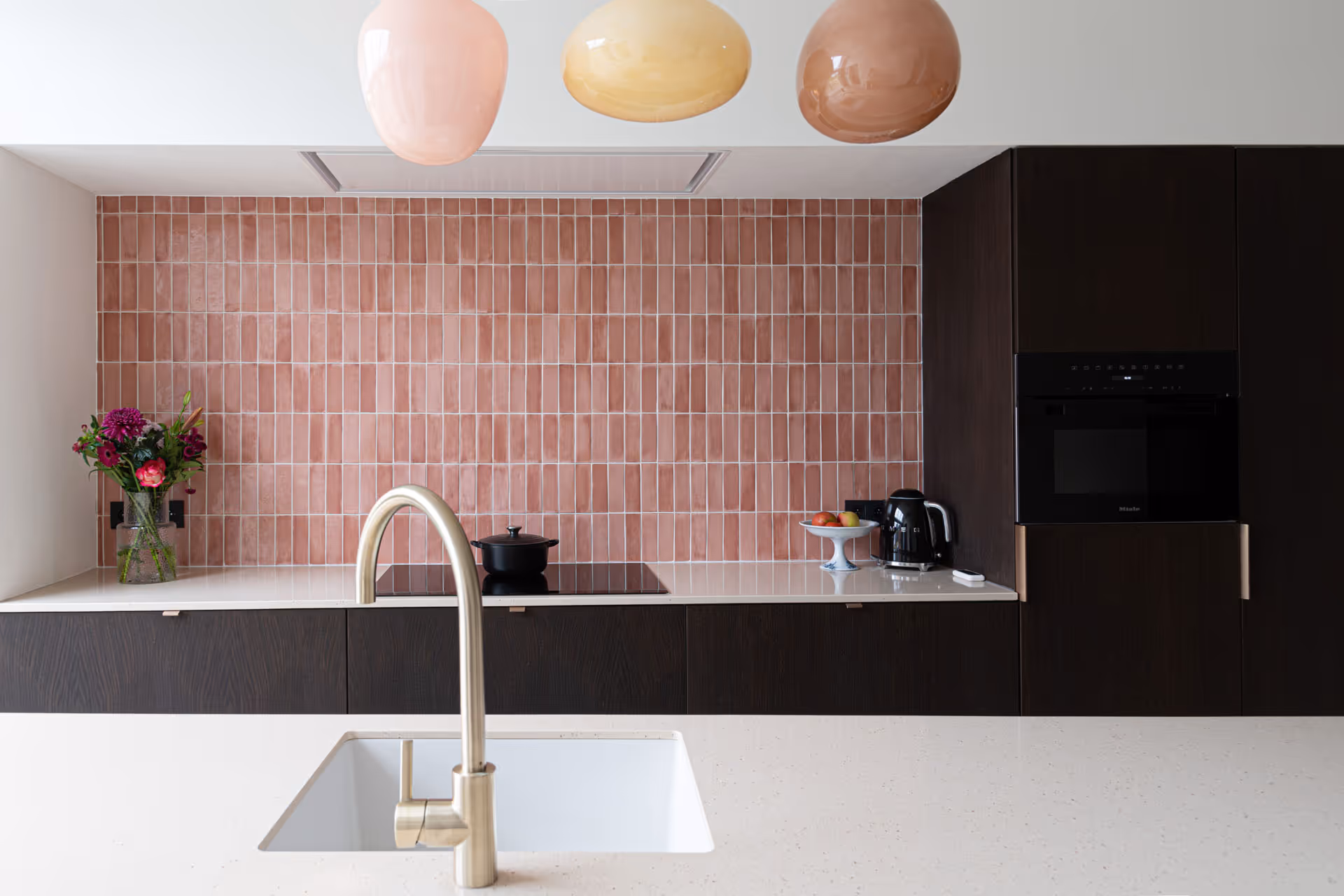 Moderne keuken met roze verticale wandtegels, zwart kookgedeelte en elegante goudkleurige kraan op wit werkblad.