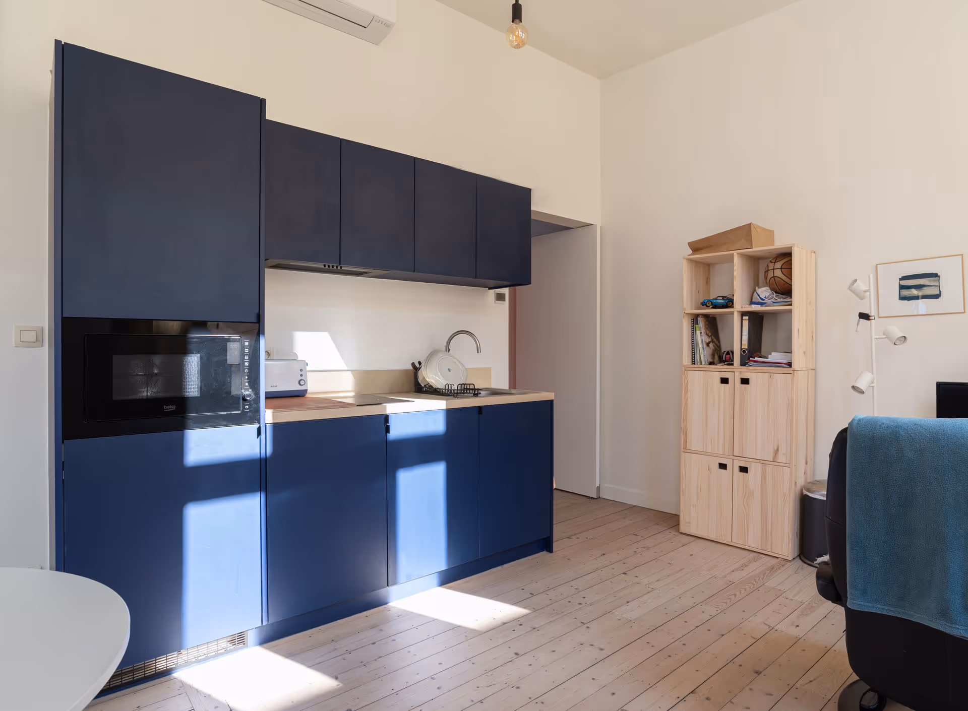 Compacte studentenkeuken met blauwe kasten, houten werkblad en licht houten vloer, geïntegreerd in een open leefruimte.