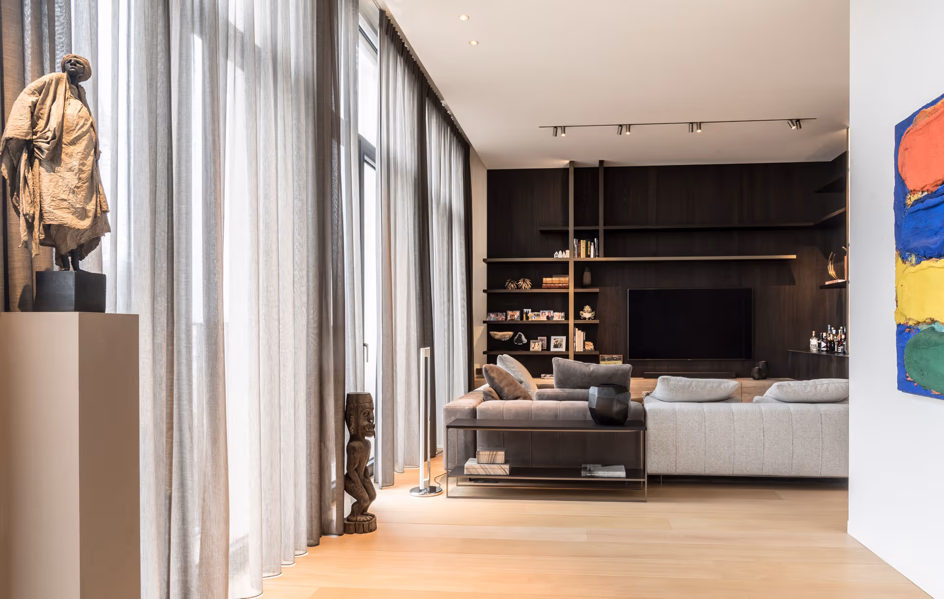 Stijlvol en modern interieur van een woonkamer met hoge ramen en lichtdoorlatende gordijnen, een strakke grijze hoekzetel, een donkere wandkast met ingebouwde tv en decoratieve kunstobjecten, waaronder sculpturen en een kleurrijk schilderij.