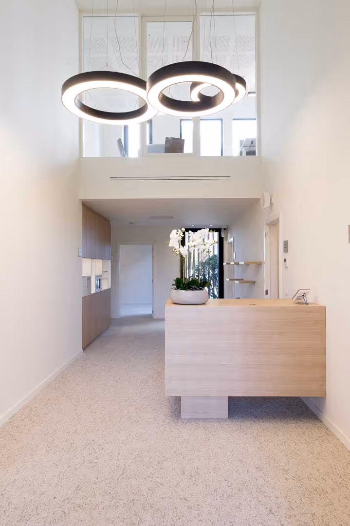 Minimalistisch en modern onthaal met een houten balie, een sierlijke orchidee in het midden, zacht tapijt en opvallende ronde designlampen aan het hoge plafond, met een glazen mezzanine die uitkijkt op de inkomruimte.