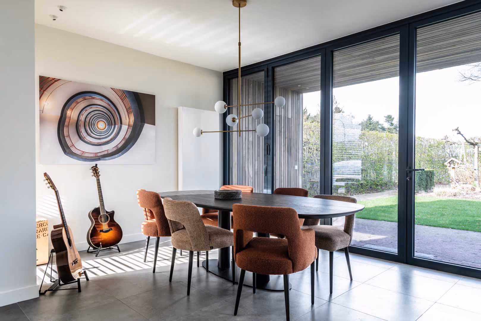 Moderne eetruimte met een ovale, donkere eettafel omringd door stoelen in warme tinten, grote raampartijen die uitzicht geven op een groene tuin, twee akoestische gitaren tegen de muur, en een opvallend spiraalvormig kunstwerk aan de muur.
