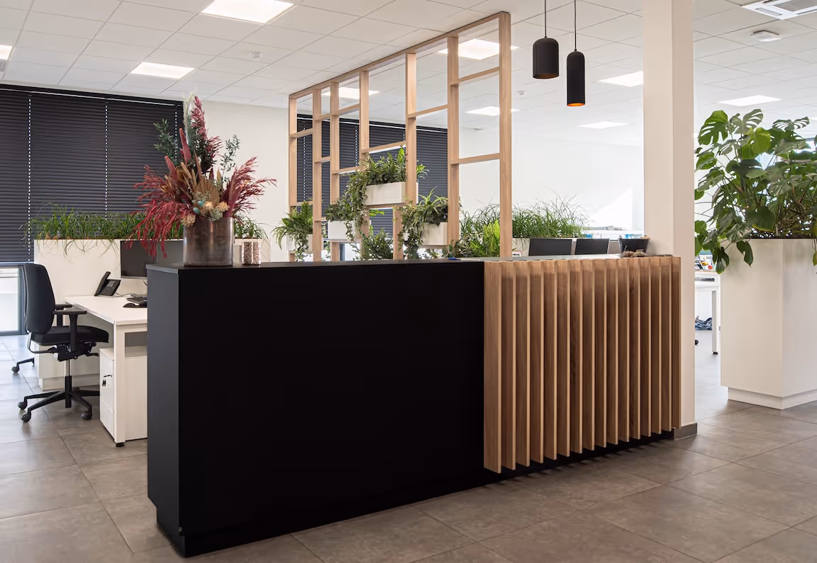 Moderne kantoorruimte met een stijlvolle zwarte ontvangstbalie met houten accenten, omringd door witte bureaus, ergonomische bureaustoelen en veel groene planten, met een houten scheidingswand die zorgt voor een natuurlijke en open sfeer.