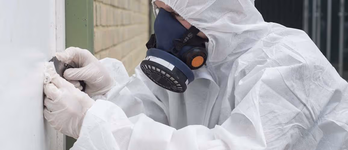 Person in protective gear scrapes paint from a wall, wearing a mask and gloves.