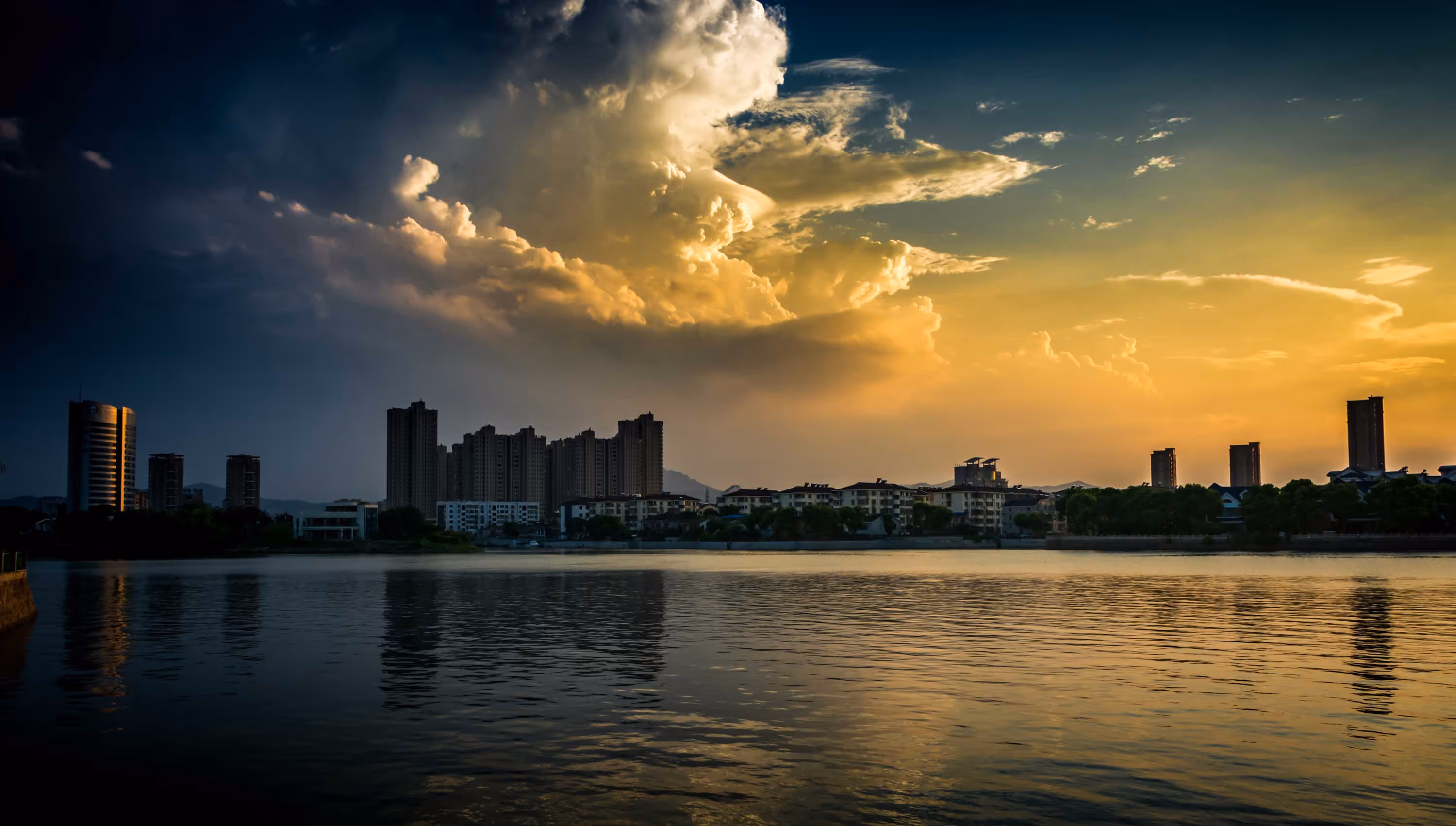 Silhouette of city buildings against a dramatic sunset sky with clouds, reflected in a calm river.