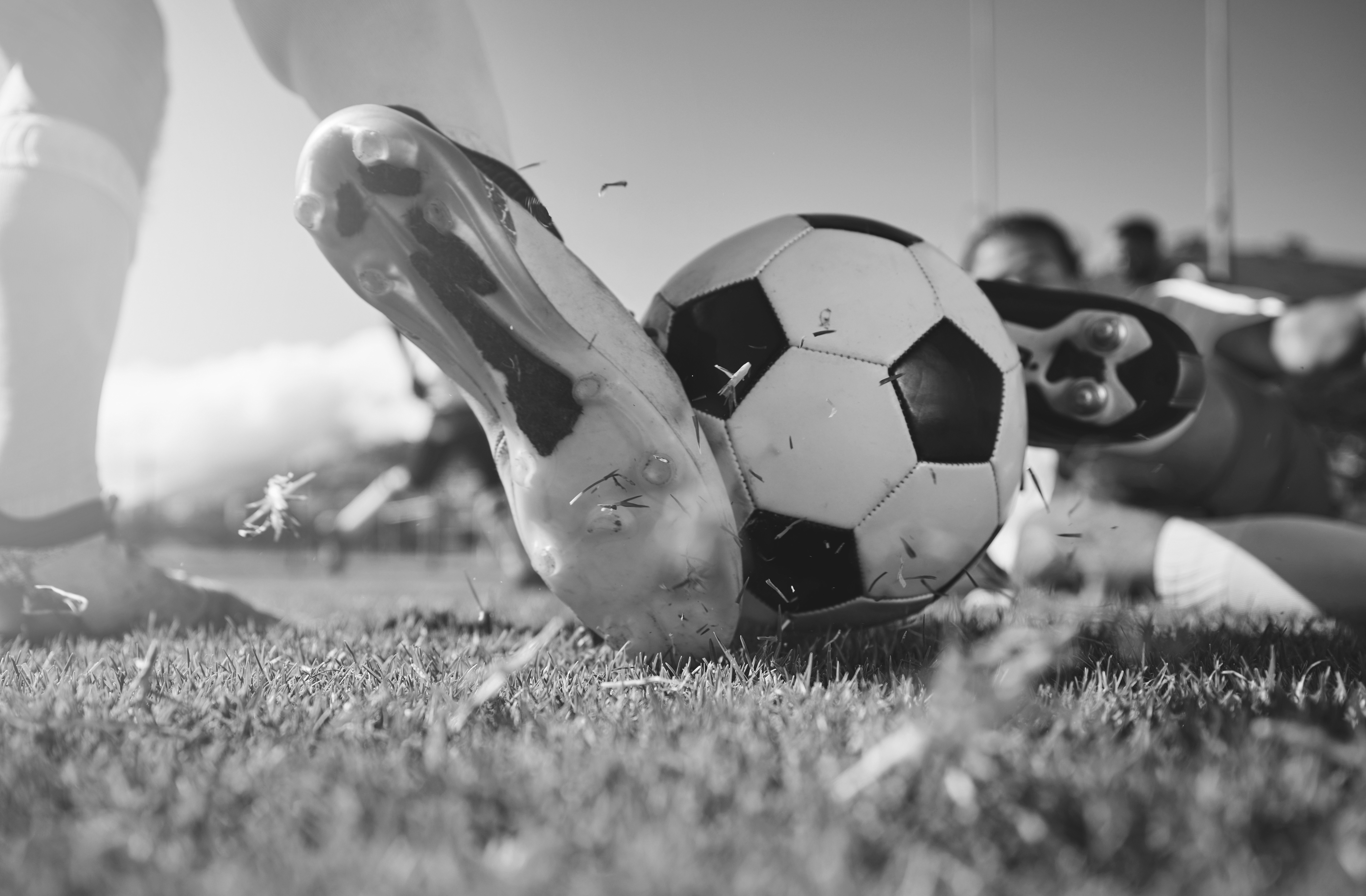 Close-up of a soccer player's cleat striking a black-and-white soccer ball on a grassy field, with an opposing player sliding in from the right.