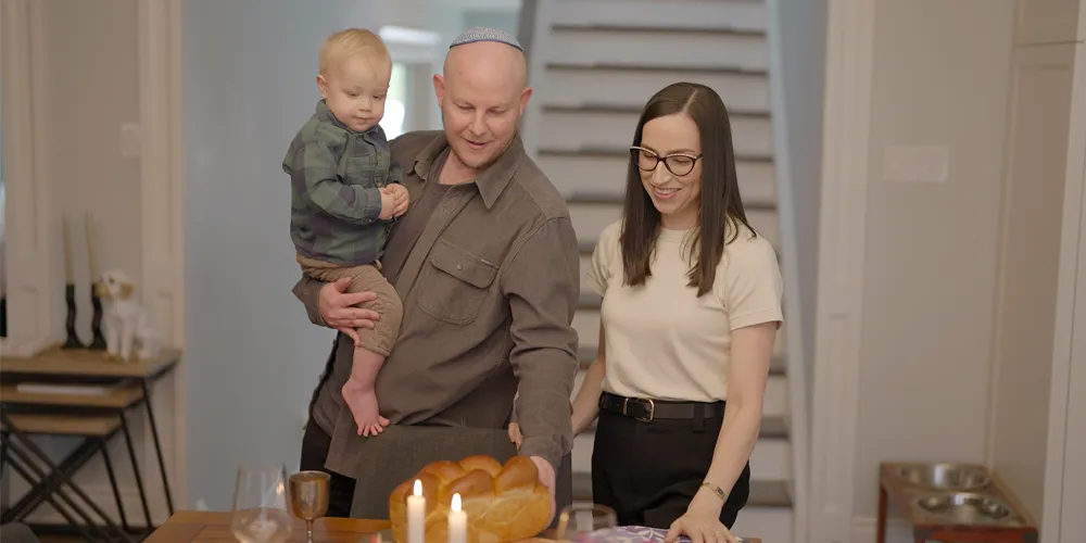 A young Jewish family including a father holding his young son in his arm while his wife stands next to him. They are celebrating Shabbat.