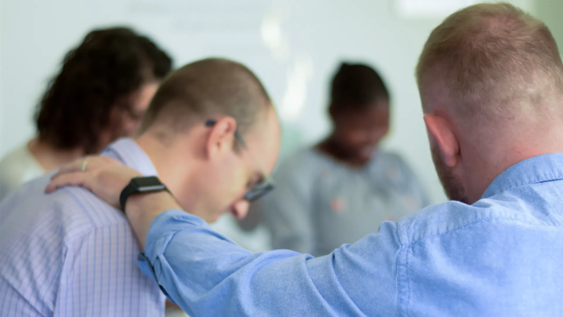 A man prays for another as he places his left arm on the other man's right shoulder. Other people are also praying in the background.