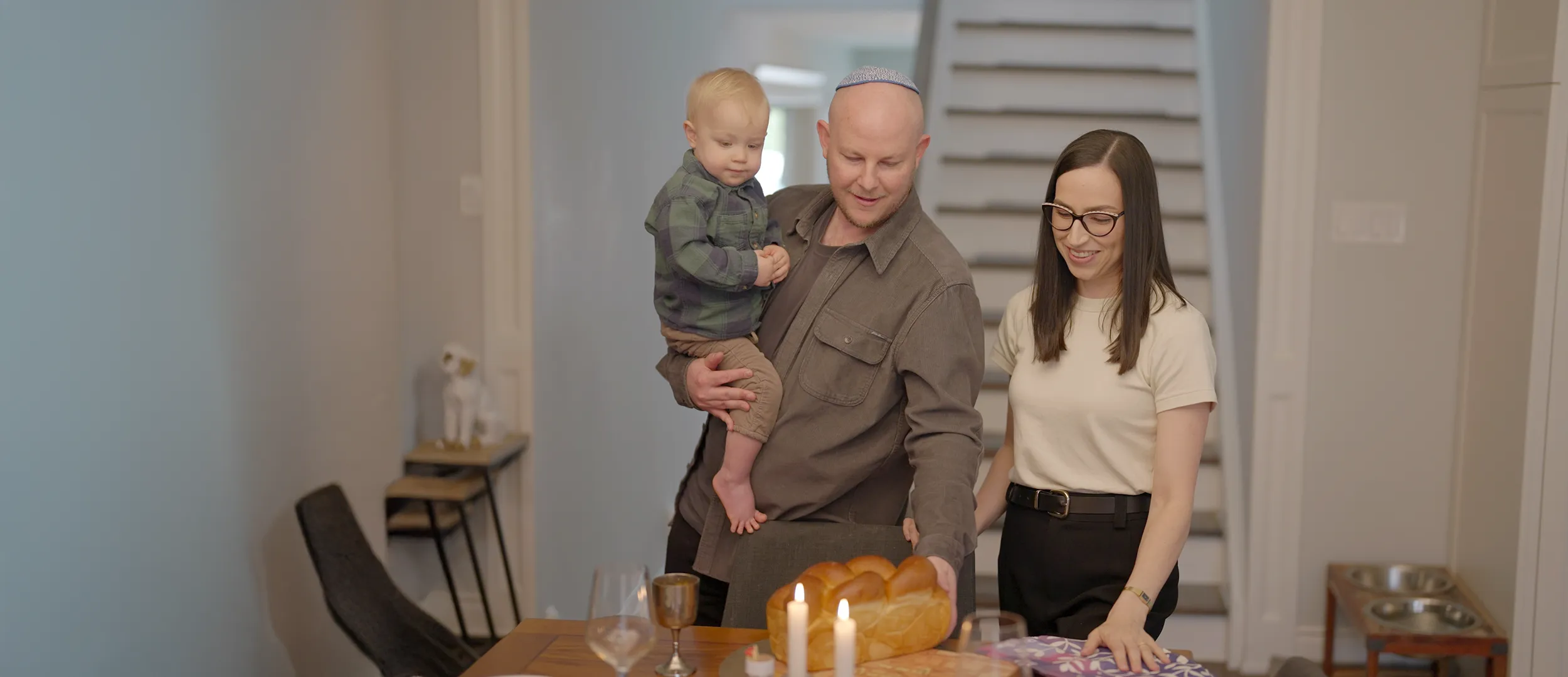 A young Jewish family including a father holding his young son in his arm while his wife stands next to him. They are celebrating Shabbat.