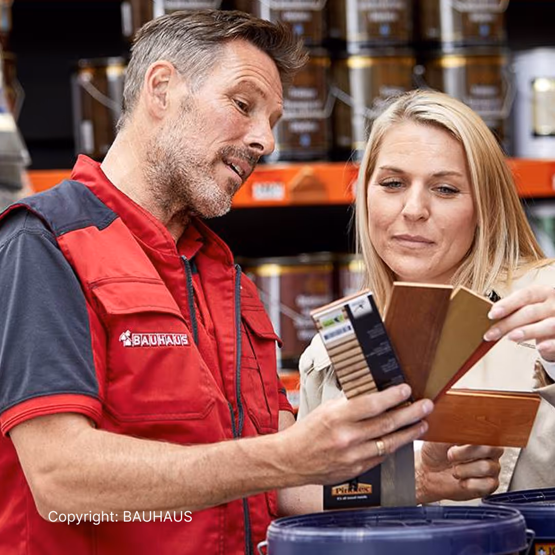 BAUHAUS store employee helping woman compare wood stain color samples in a hardware store aisle.