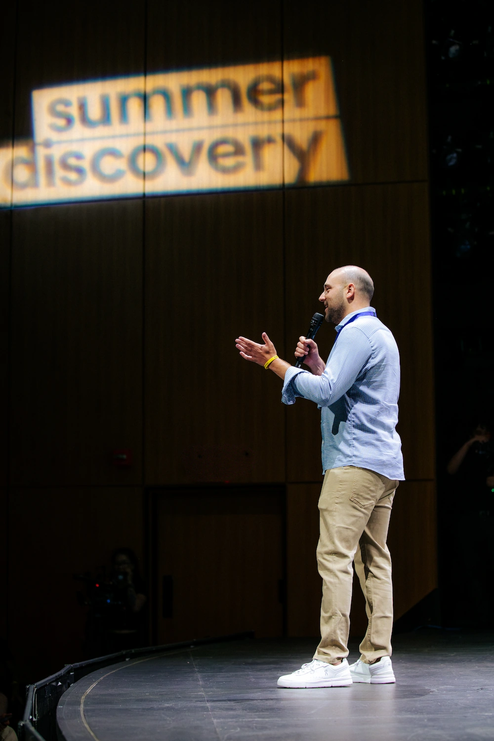 Man in light shirt and khaki pants speaking with microphone on stage under summer discovery sign.