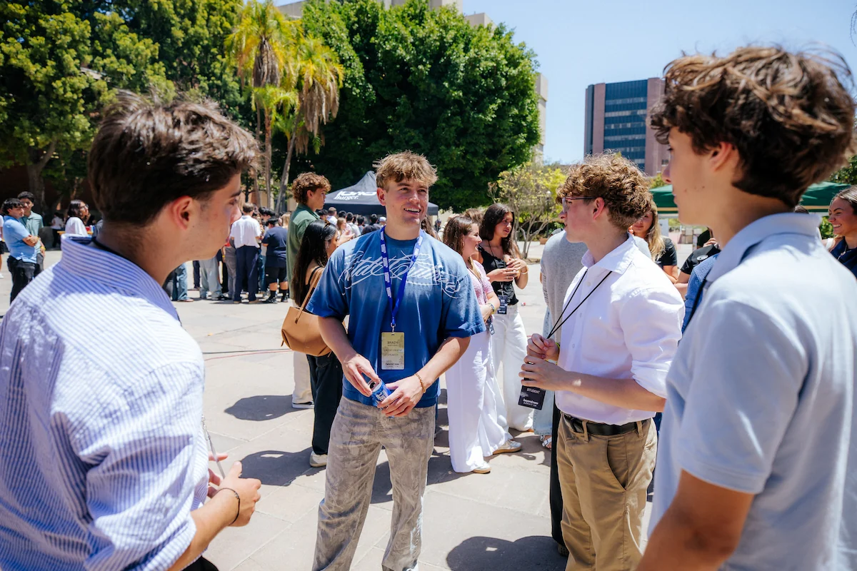 Group of young people talking outdoors in a sunny urban park with trees and buildings in background.