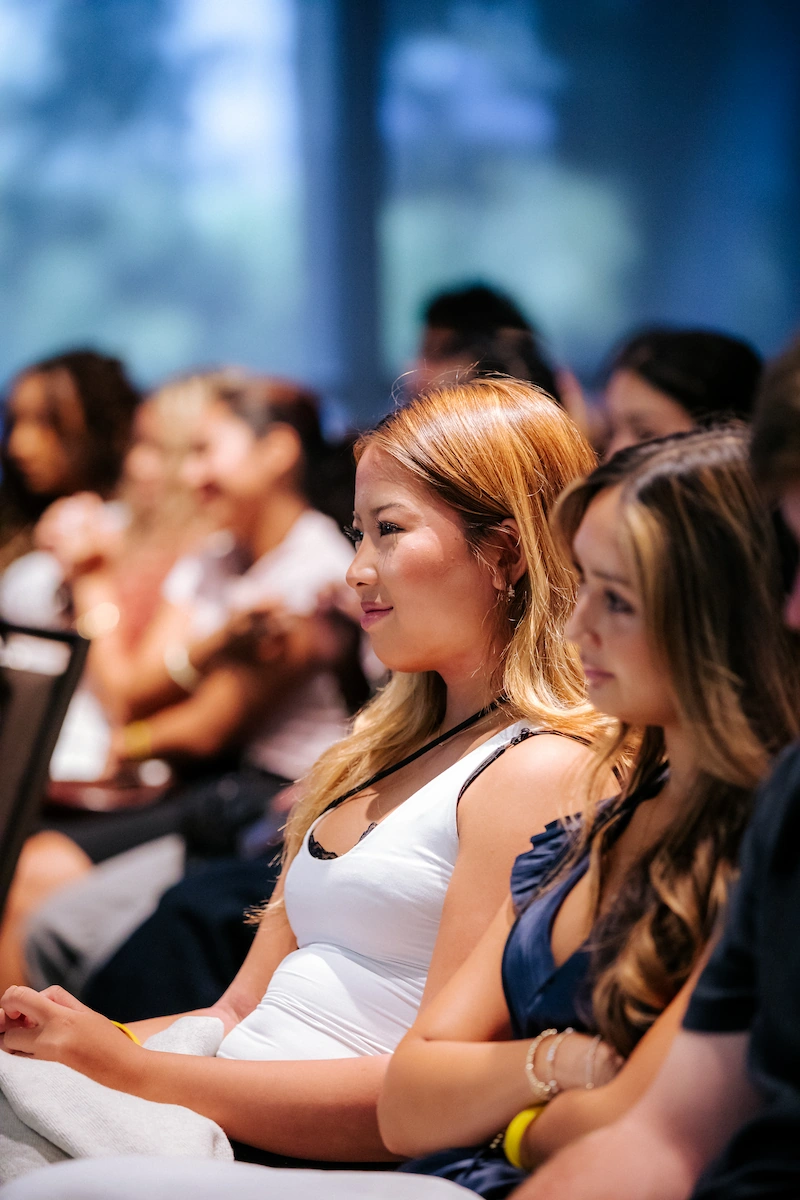 Women seated in a row attentively listening at an indoor event with blurred background attendees.