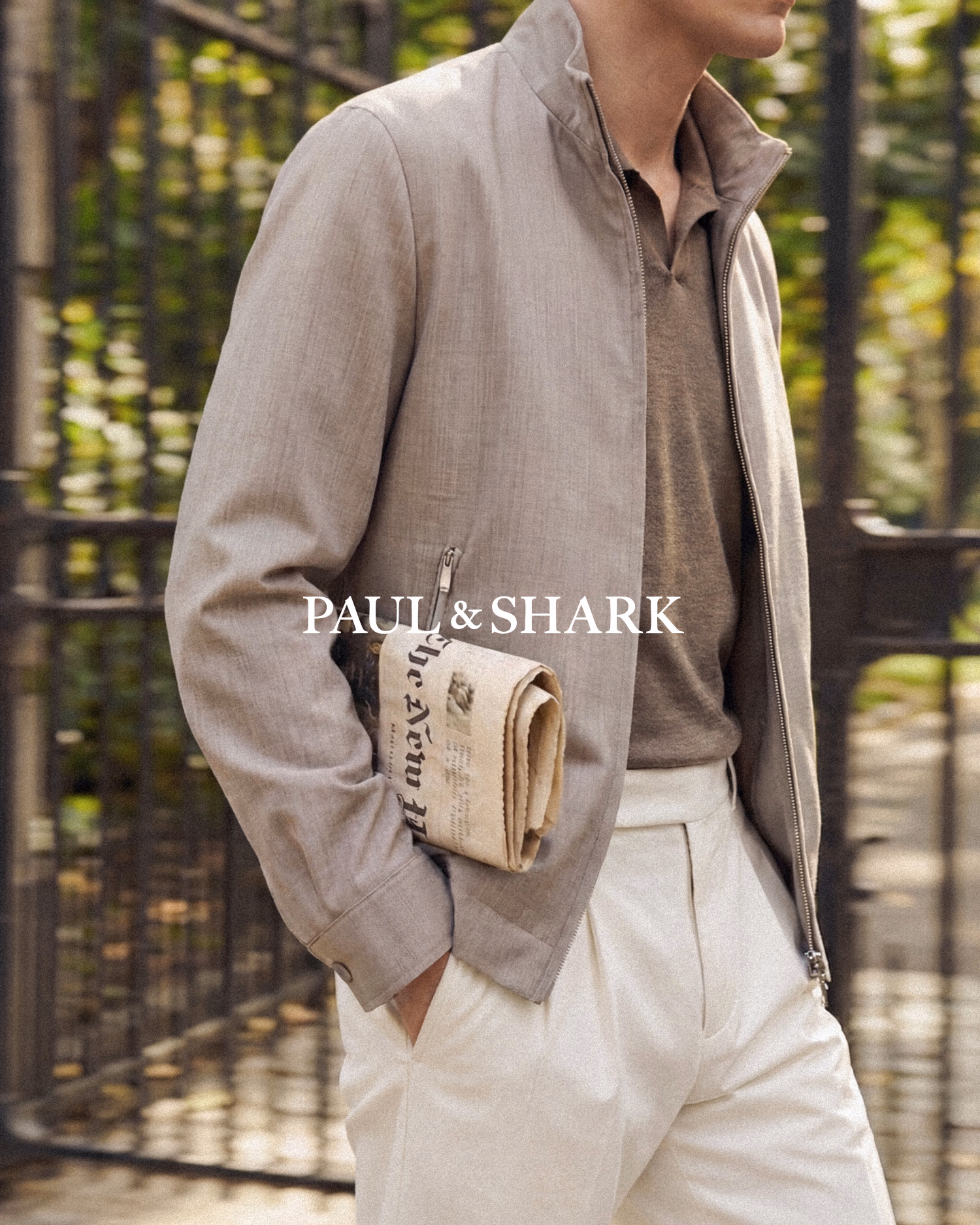 Paul & Shark FW26: man in a taupe zip jacket and cream trousers with a newspaper clutch