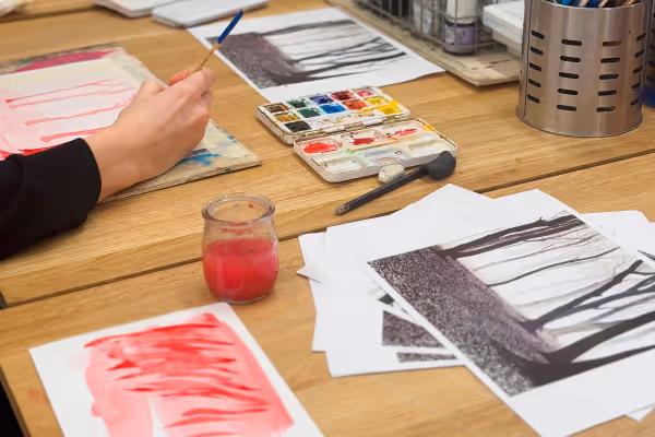 Un élève peint une aquarelle sur une table d'atelier. Gros plan sur la main qui fait un lavis. Sur la table de dessin sont disposés du matériel pour réaliser la peinture.