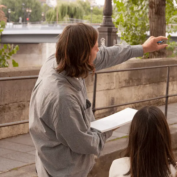 Un artiste-enseignant montre une proportion à l'aide de l'empan de sa main pendant un cours de carnet de voyage en extérieur à Paris sur les quais de Seine.