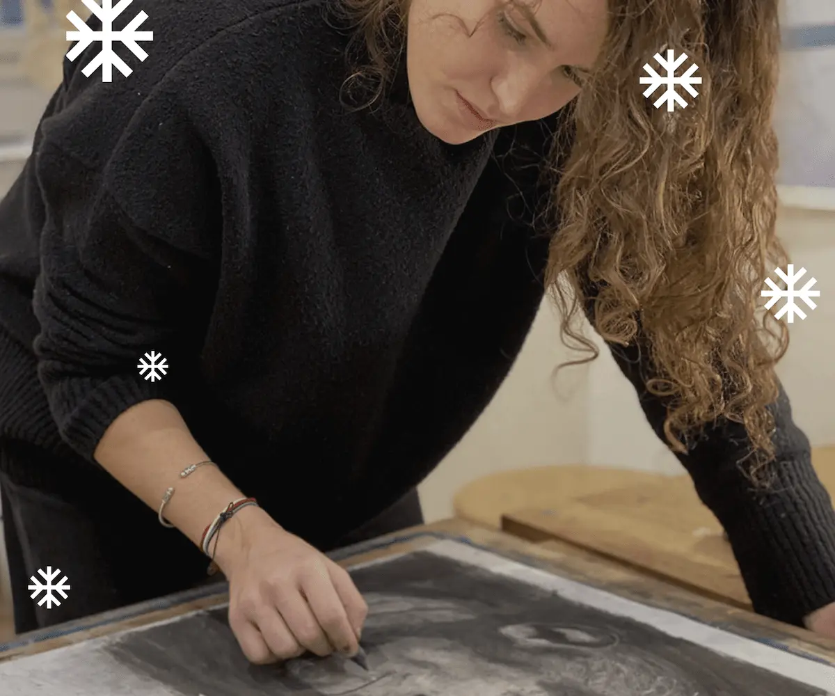 Une personne dessine avec un stylo fin sur une grande feuille blanche avec des taches colorées à l'aquarelle, à côté d'une palette de peinture et d'un carnet jaune.