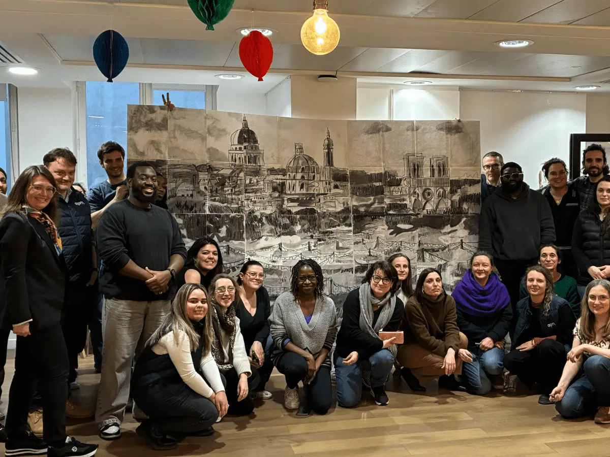 Une personne dessine avec un stylo fin sur une grande feuille blanche avec des taches colorées à l'aquarelle, à côté d'une palette de peinture et d'un carnet jaune.