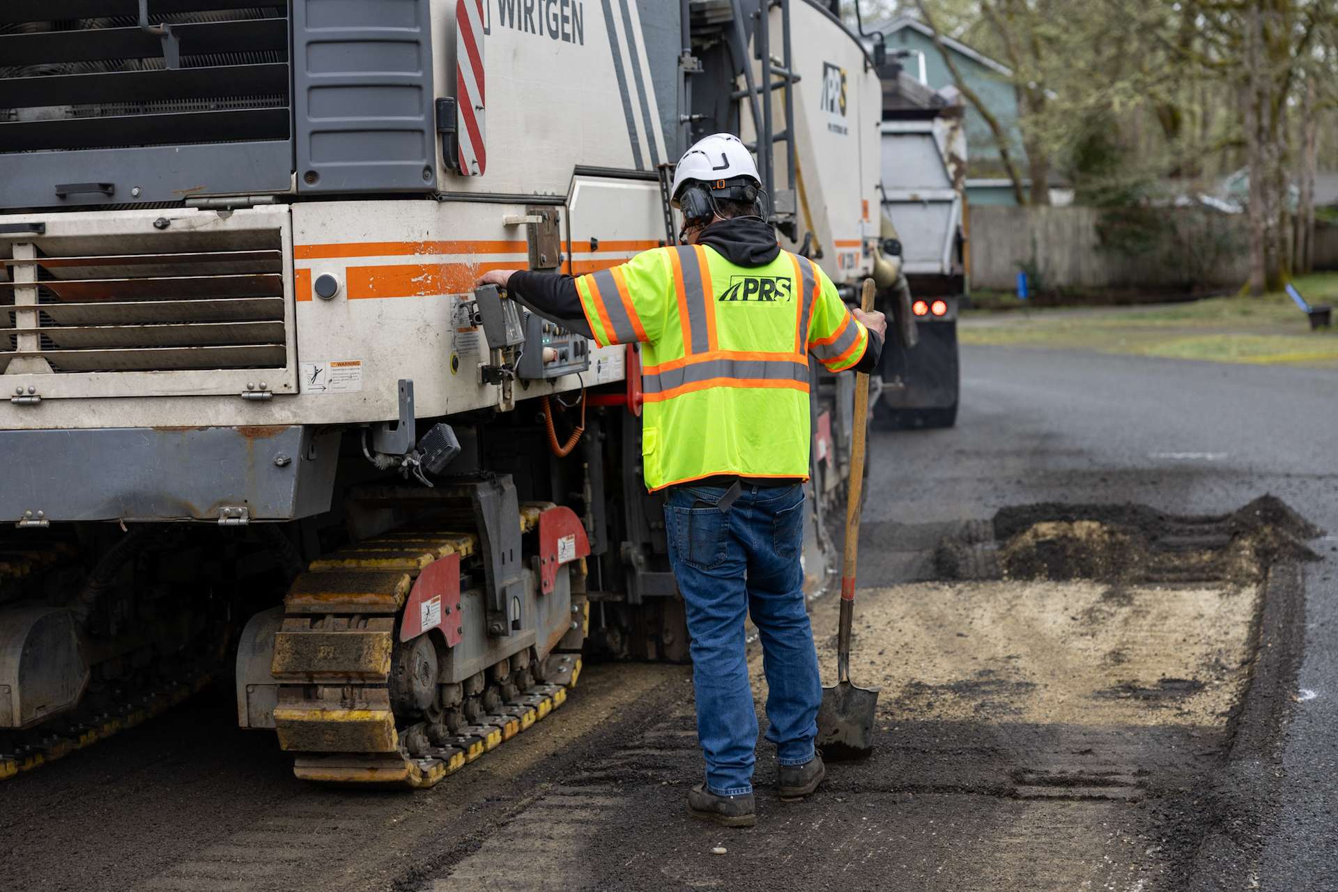 PR Systems operator controls Wirtgen milling machine during precise asphalt removal on city street.