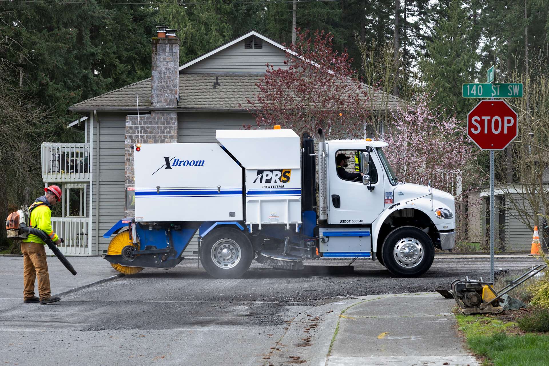PR Systems street sweeper truck cleaning up milled asphalt as a worker uses a leaf blower for fine debris.