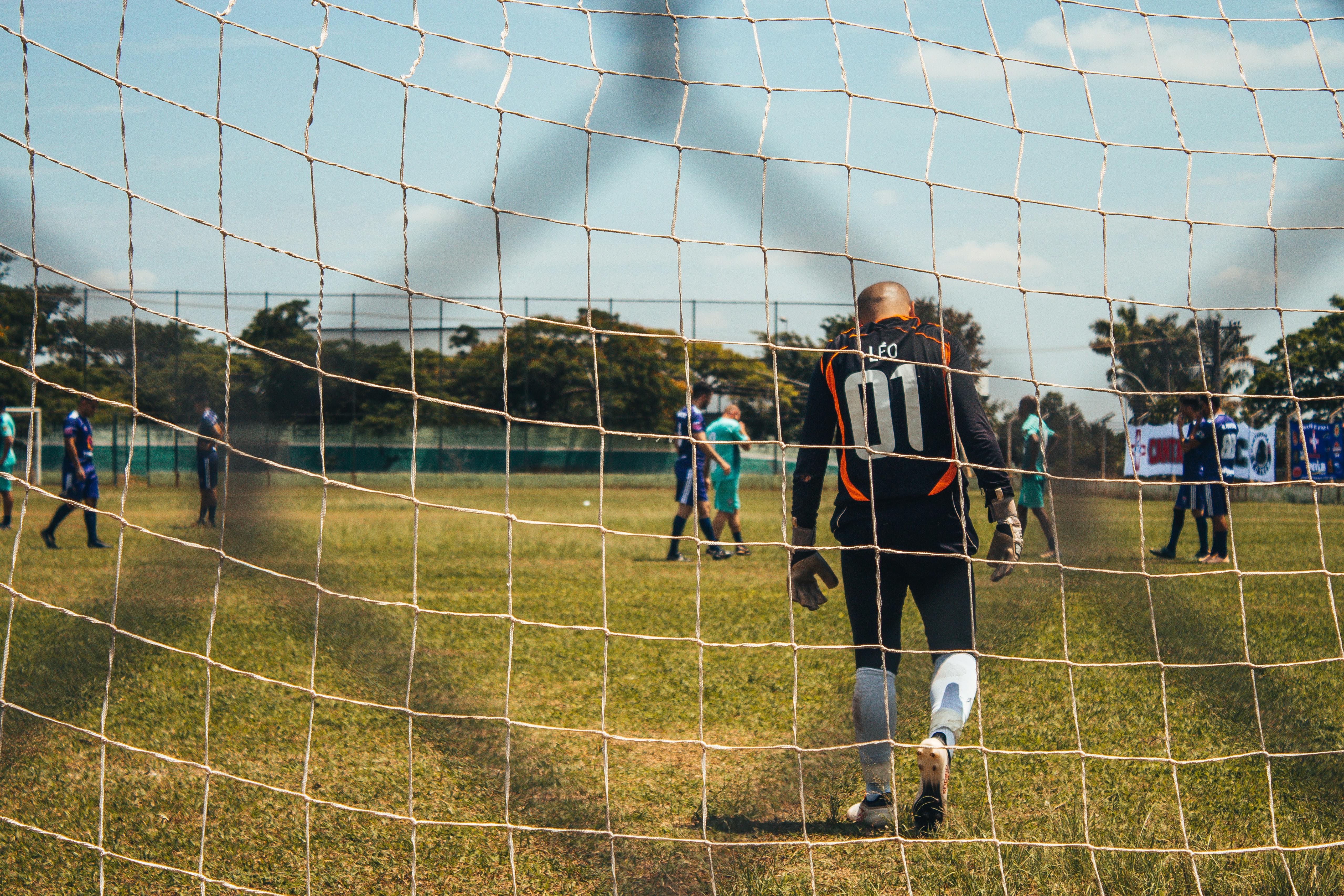 Portero de fútbol con uniforme negro y naranja con el número 01 en la espalda, de espaldas dentro de la portería en un campo de césped, con jugadores y árboles al fondo.