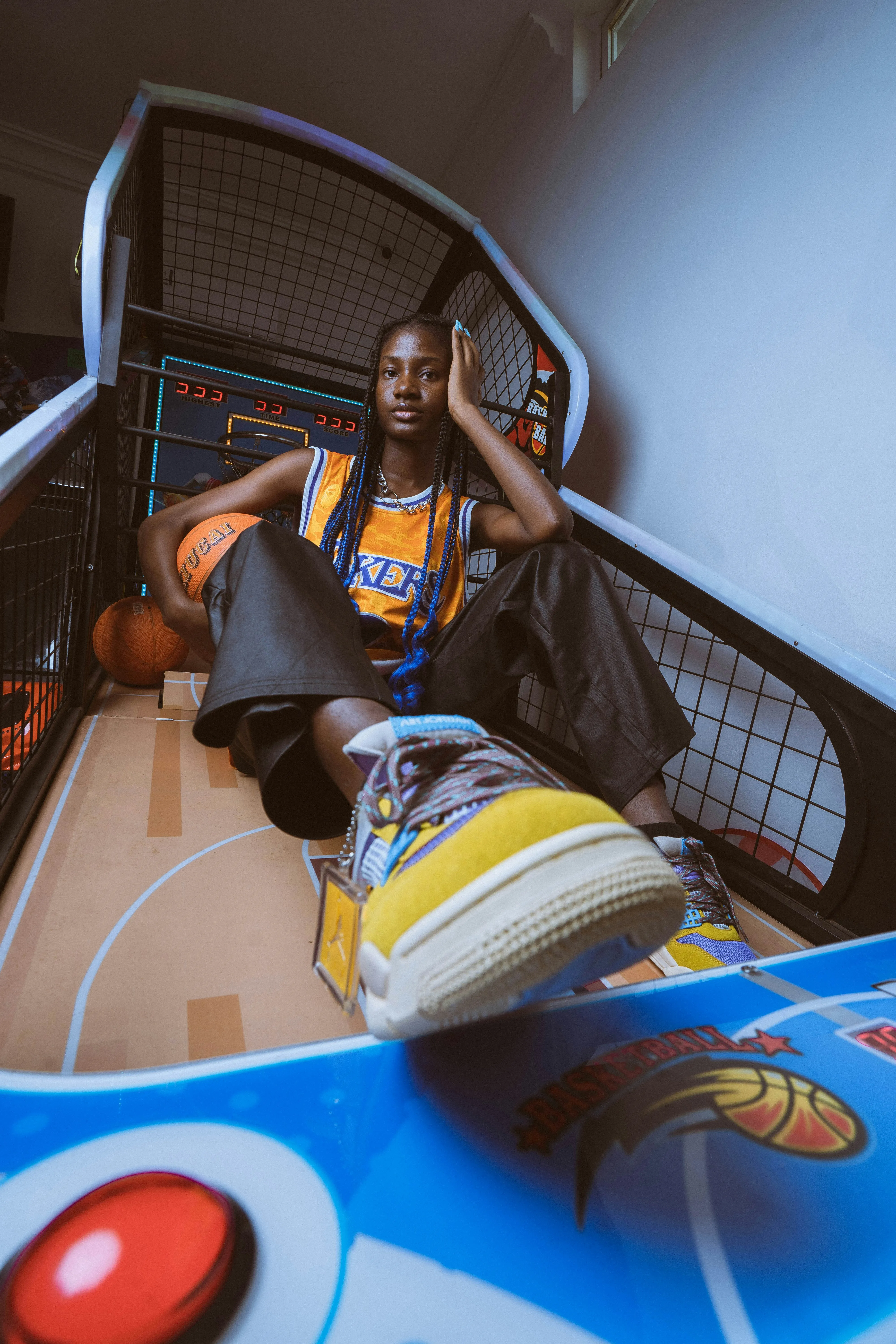 Joven con camiseta de Lakers y trenzas azules, sentada en una máquina de baloncesto arcade con una bola de baloncesto.