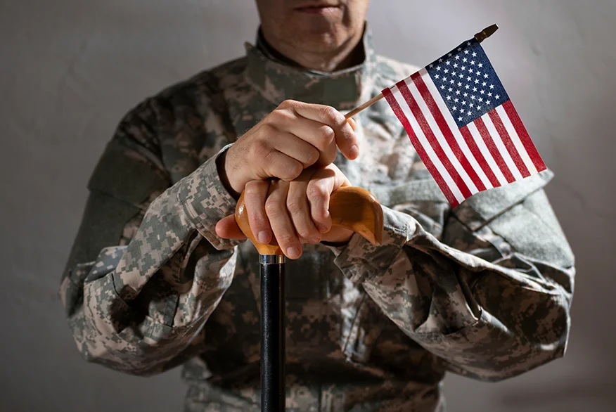 Soldado en uniforme de camuflaje sosteniendo un bastón y una bandera estadounidense que simboliza el apoyo y la resistencia de los veteranos