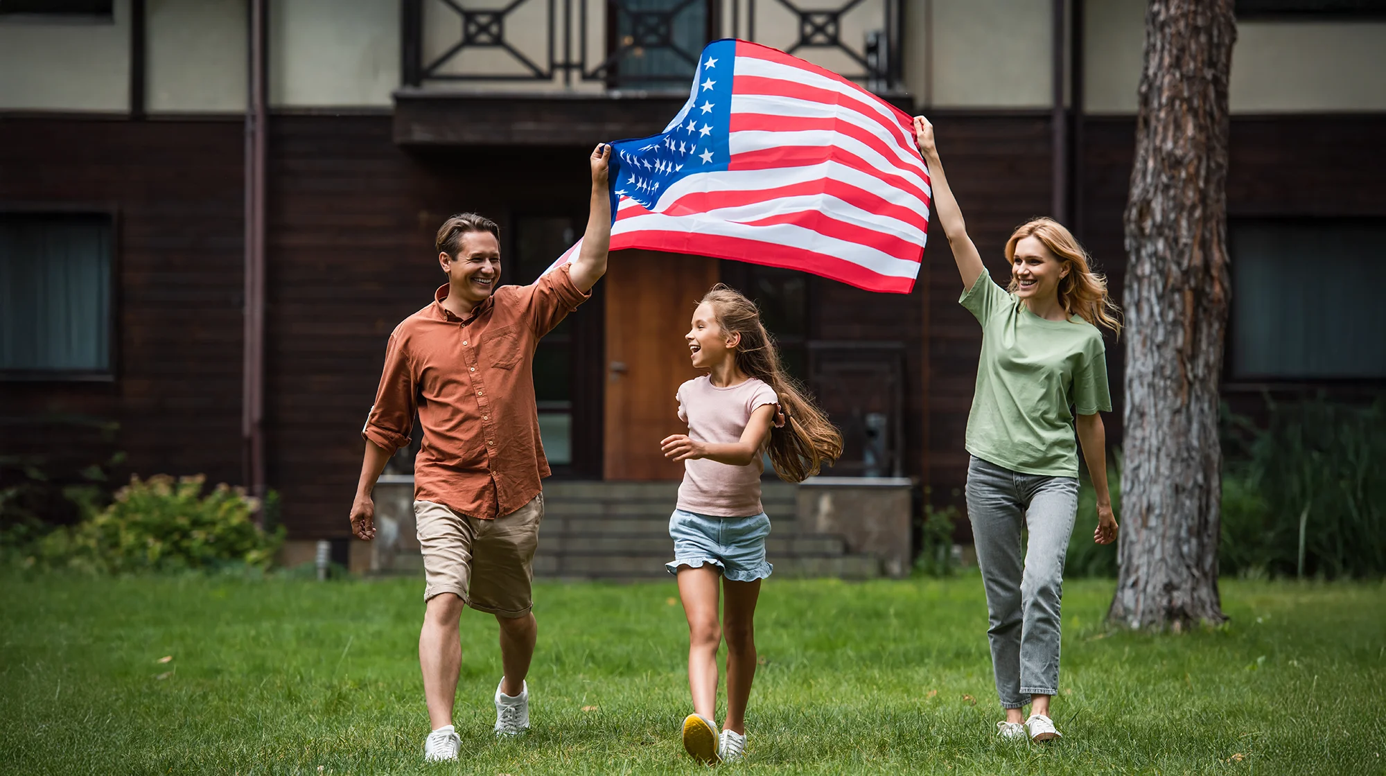 A happy family holding a US flag waking on the grass in front of their house.