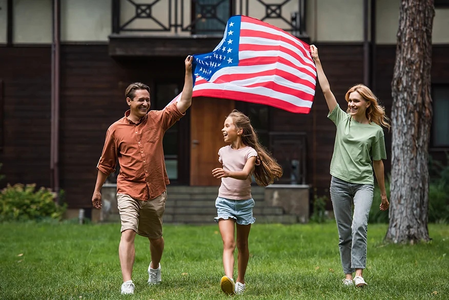 A happy family holding a US flag waking on the grass in front of their house.