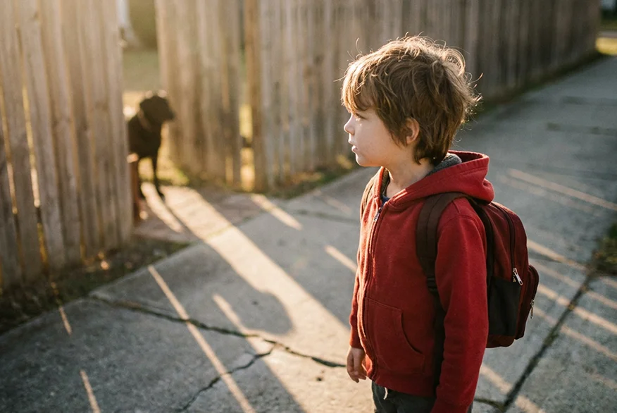 Child in a red hoodie with a backpack standing on a sunlit sidewalk, a dog by a wooden fence in the background.